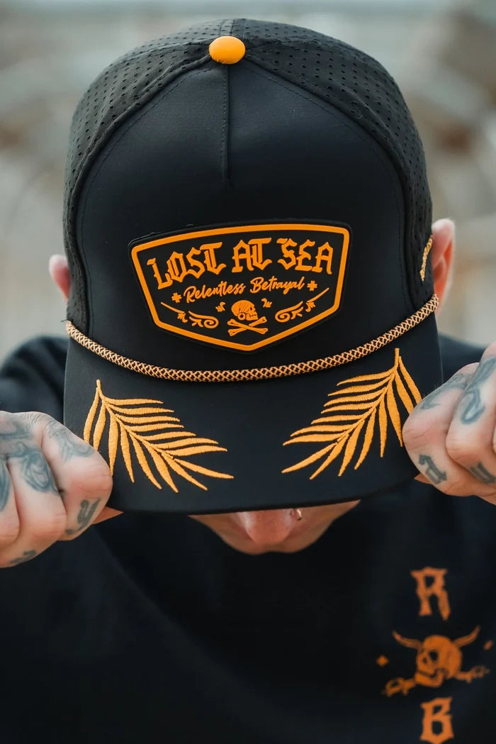 Wearing the Lost At Sea PVC Snapback by Relentless Betrayal, a person sports a black cap with vibrant orange Lost at Sea text. The brim features orange palm leaves, while tattooed hands grip the cap and a lip piercing adds to their pirate vibe.