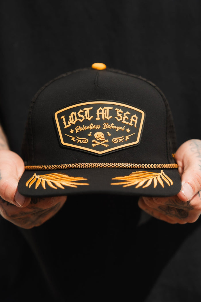 A person holds a Lost At Sea PVC Snapback by Relentless Betrayal, featuring gold lettering and a brim adorned with gold leaf embroidery. This water-resistant black cap is the perfect mix of durability and style for a pirate lifestyle against a dark, neutral backdrop.