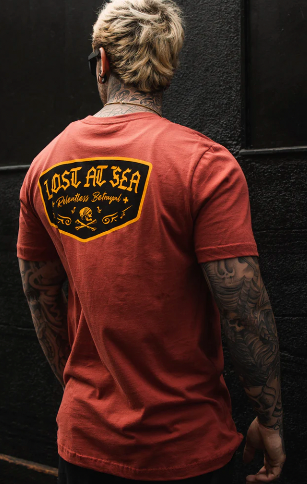 A tattooed person with blonde hair stands against a dark wall, wearing the Lost at Sea Salmon Tee by Relentless Betrayal. The fine jersey cotton T-shirt features Lost At Sea Relentless Betrayal and a skull graphic on the back.