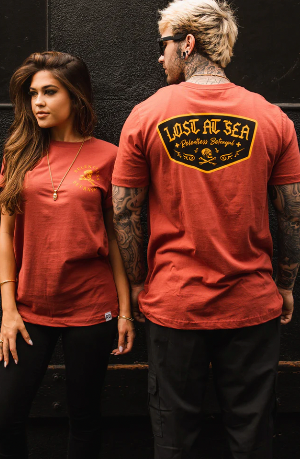 A woman and man stand against a dark wall, wearing red Lost at Sea Salmon Tees by Relentless Betrayal, made from fine jersey cotton. She faces forward, showing a small chest logo, while he turns to display a large back graphic. Both flaunt intricate tattoos.