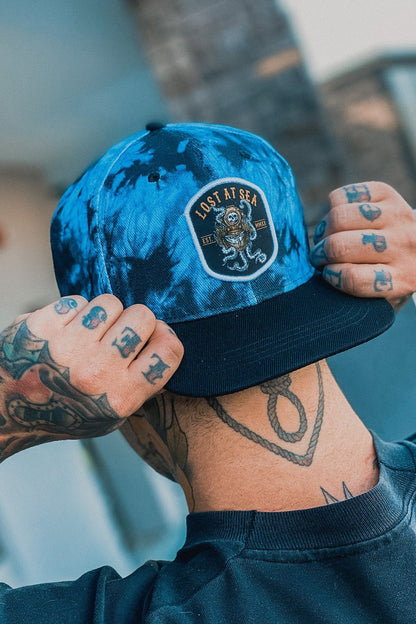 A person with tattooed hands adjusts a Relentless Betrayal Lost At Sea Snapback - Tie Dye, featuring an octopus design and the words LOST AT SEA. Wearing a black shirt, they stand against a slightly blurred backdrop of part of a building.