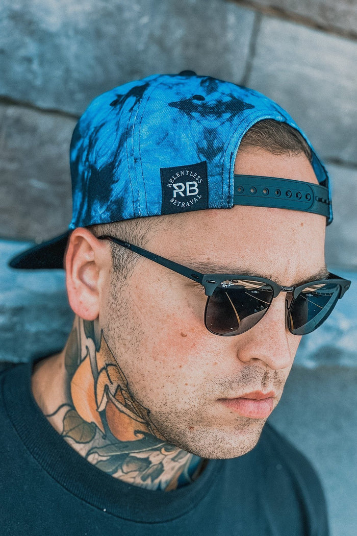 A man wears a Lost At Sea Snapback - Tie Dye by Relentless Betrayal backwards with sunglasses. A tattoo adorns his neck above a black shirt, against stone-textured wall panels.