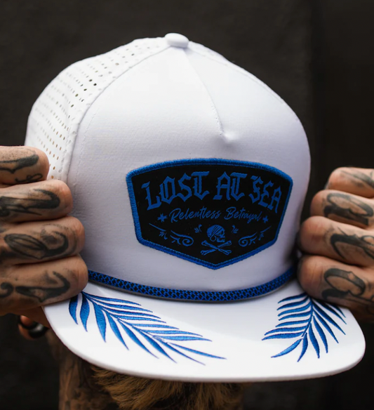 A person with tattooed hands holds the Lost at Sea Whiteout Snapback by Relentless Betrayal. The hat features a blue and black Lost At Sea: Relentless Betrayal patch and a brim printed with blue palm leaves, enhancing its nautical charm.