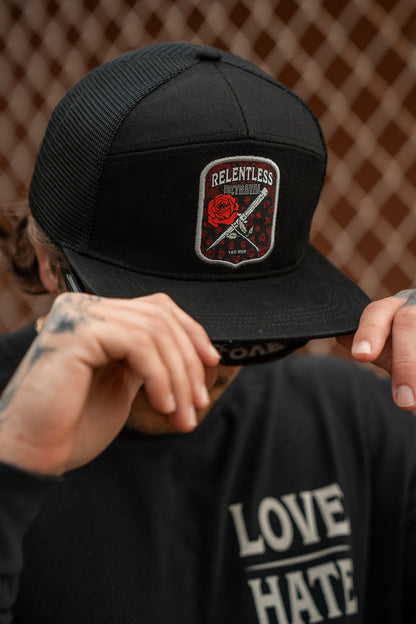 A person adjusts a black snapback from Relentless Betrayal featuring the Love/Hate 7 Panel design, with a Relentless patch showing a red rose and crossed chains. Tattooed hands adjust it, while they sport a shirt with Love and Hate. A chain-link fence is in the background.