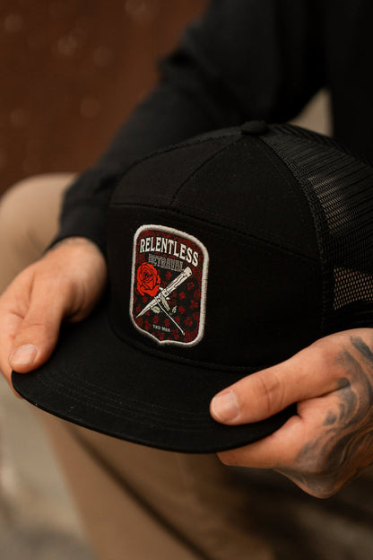 A tattooed individual holds a handmade black Love/Hate 7 Panel Trucker Snapback by Relentless Betrayal, adorned with a rose and two swords patch. Theyre wearing a black long-sleeve shirt and beige pants.
