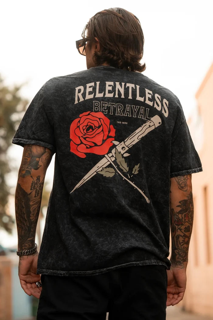 A person in the Love/Hate Premium T-Shirt, featuring a red rose and crossed knife design with Relentless Betrayal above, showcases a love/hate vibe. The unisex jersey cotton shirt accents their long hair and tattooed arms.