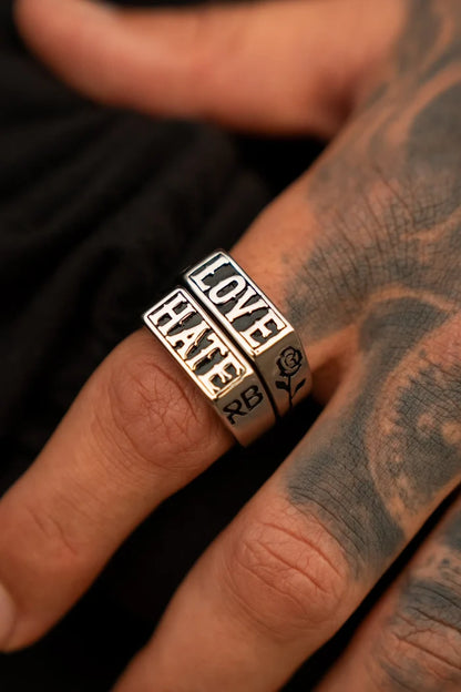 A hand showcases a duo from Relentless Betrayals Love/Hate Rings on the middle finger—one with LOVE and one with HATE, both in stainless steel. Detailed tattoos, like a rose, add artistry against a stark black backdrop.
