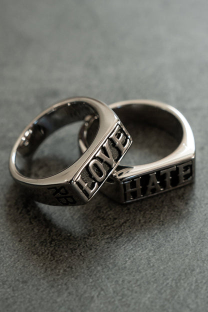 Two silver rings lie on a gray surface: one is engraved with LOVE and the other with HATE. This handcrafted designer duo, from Relentless Betrayals Love/Hate Rings collection, forms an overlapping design.