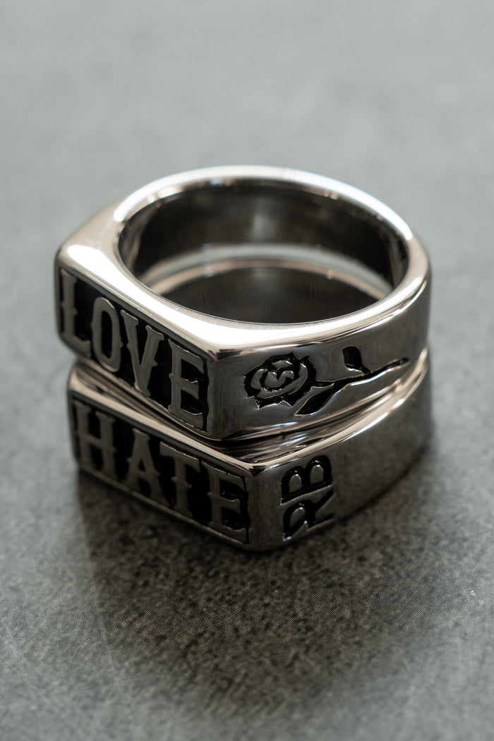 The Love/Hate Rings by Relentless Betrayal are a stunning duo stacked on a gray surface. The top stainless steel piece is engraved with LOVE alongside a rose design, while the bottom reads HATE, each telling its own handcrafted jewelry tale.