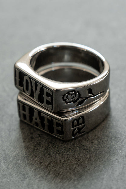 The Love/Hate Rings by Relentless Betrayal are a stunning duo stacked on a gray surface. The top stainless steel piece is engraved with LOVE alongside a rose design, while the bottom reads HATE, each telling its own handcrafted jewelry tale.