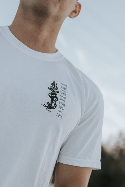 A person wears the 13th brand Lucifers Demise Premium Tee- White, featuring a black dragon design and text, against a sky backdrop. Captured from a low angle, the photo beautifully highlights the casual outfit and serene setting.