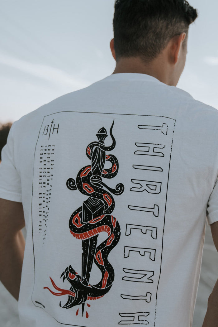 A person outdoors dons a white Lucifers Demise Premium Tee by 13th, featuring a graphic of a sword with an entwined snake symbolizing Crush Satan, and the word THIRTEENTH vertically along the side, set against a serene clear sky backdrop.
