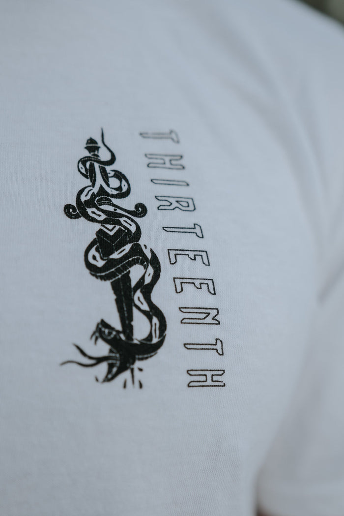 Close-up of the Lucifers Demise Premium Tee- White by 13th, displaying a black design with intertwined snakes and symbols beside the vertical word THIRTEENTH, subtly embodying the Grace of Our Lord against a blurred background.