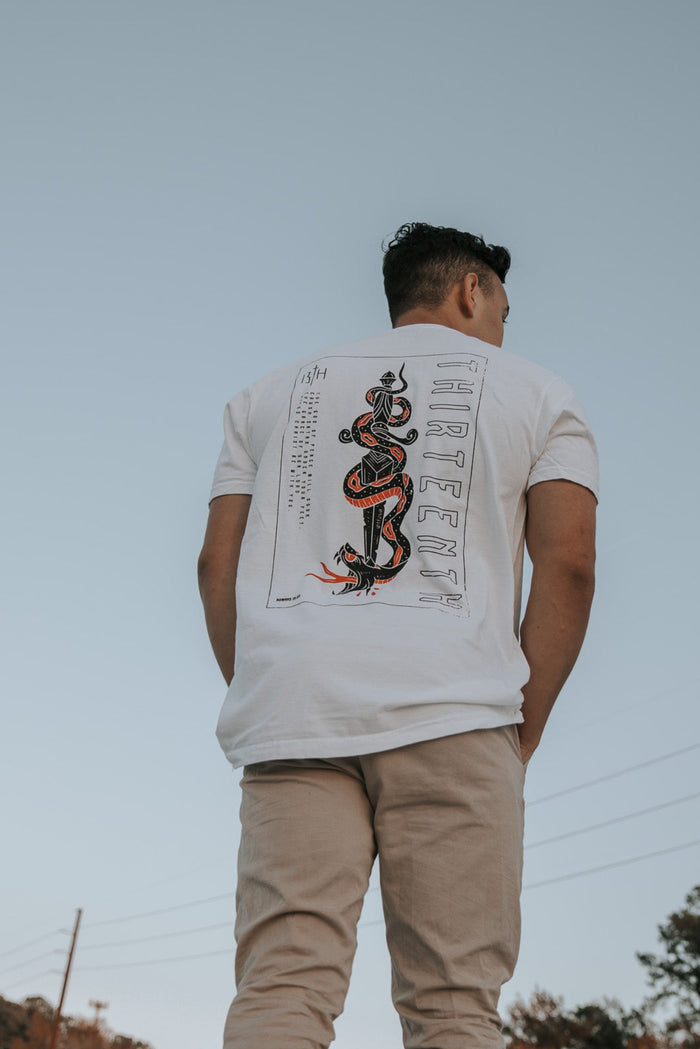A man wearing the 13ths Lucifers Demise Premium Tee- White with a snake and Crush Satan graphic stands outdoors in beige pants, set against a clear sky with trees and power lines providing a serene background.