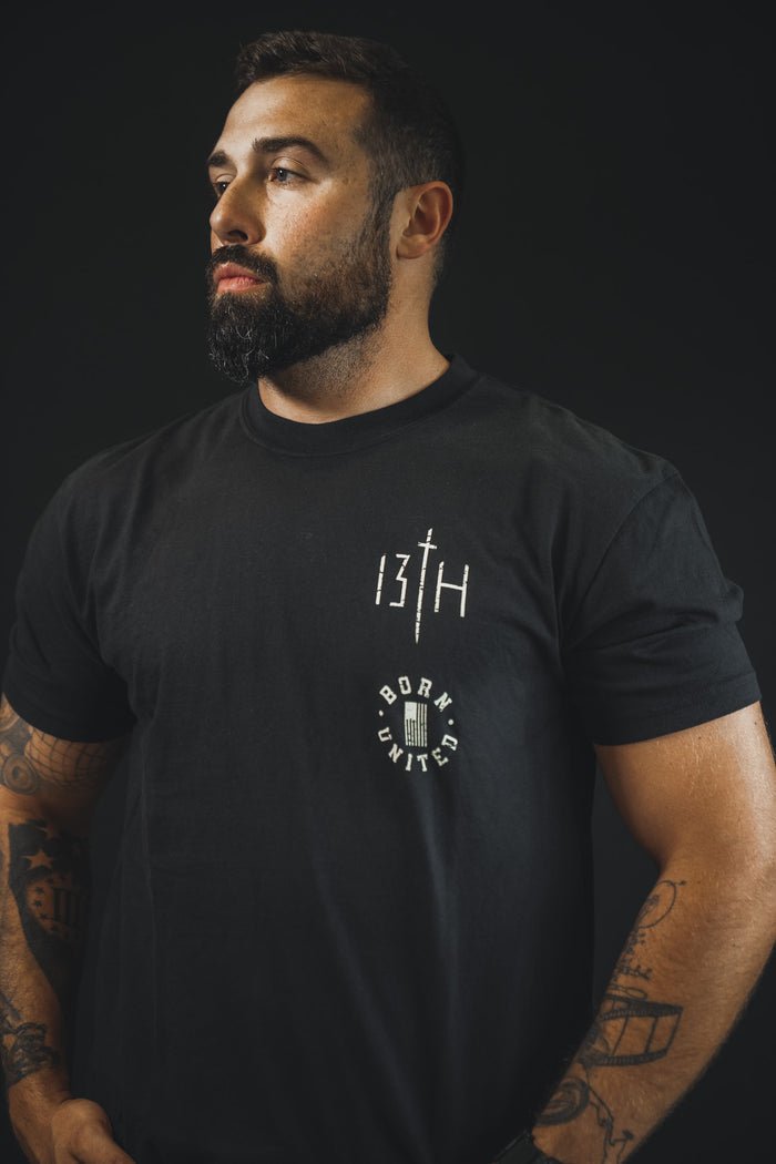 A bearded, tattooed man wears the “Make No Peace” Premium Tee by Born United Private Label, featuring bold statement graphics, against a dark background.