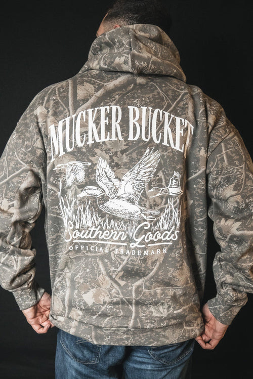 A person wears a Mallard Deadwood Camo Hoodie by Mucker Bucket—featuring relaxed fit and illustrated ducks in flight—standing against a plain black background.