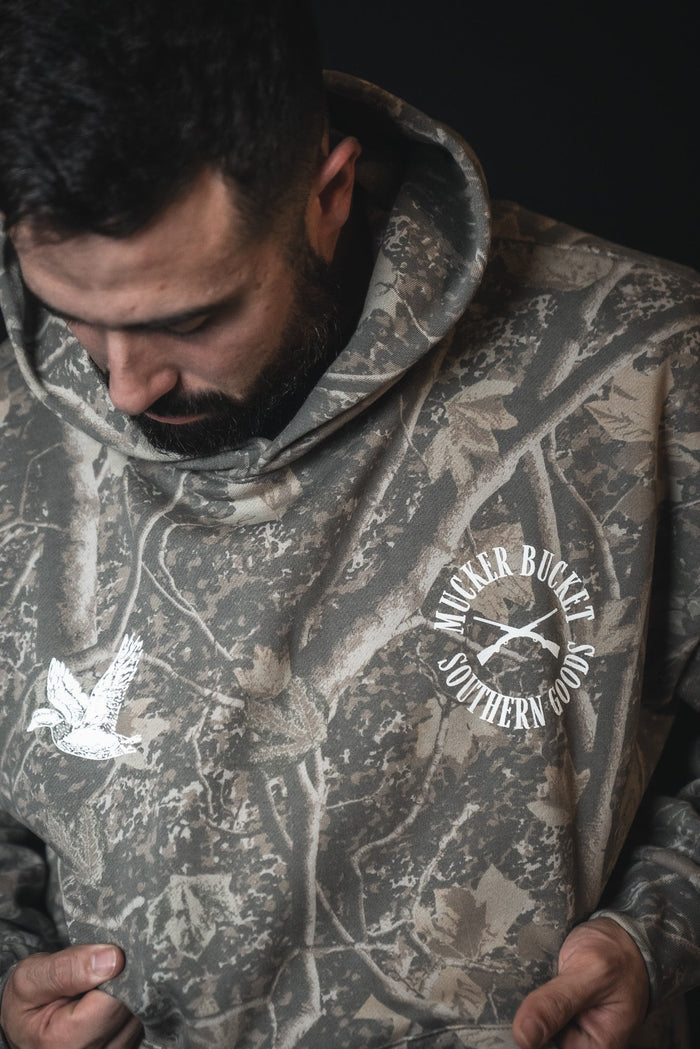 A bearded man looks down, wearing the Mallard Deadwood Camo Hoodie from Mucker Bucket, featuring a flying duck graphic. He holds the sides of this comfortable hoodie for a relaxed fit.