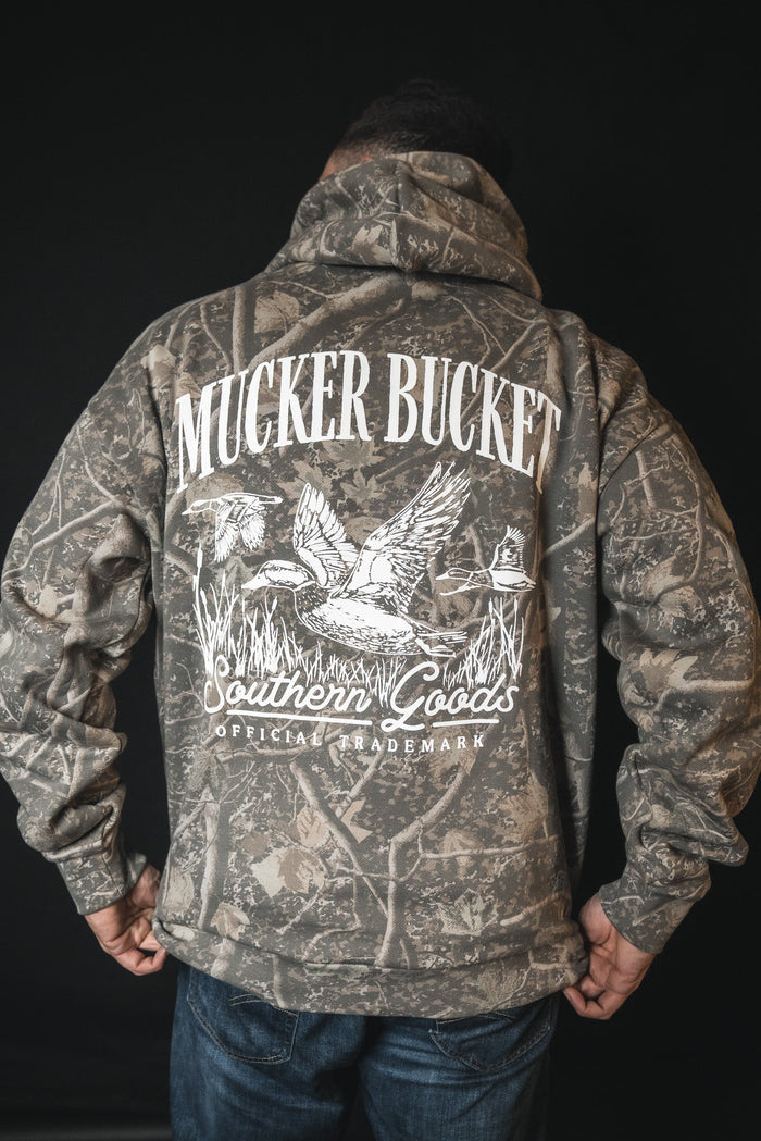A person wearing the Mallard Deadwood Camo Hoodie by Mucker Bucket, featuring flying ducks and white Southern Goods Official Trademark print on the back, stands against a black background.