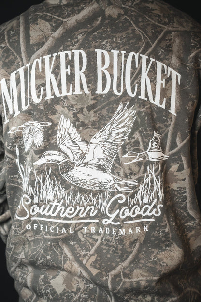 Someone wears the Mallard Deadwood Camo Hoodie by Mucker Bucket, featuring white MUCKER BUCKET text above a graphic of three ducks with grass, and "Southern Goods Official Trademark" printed below the image.