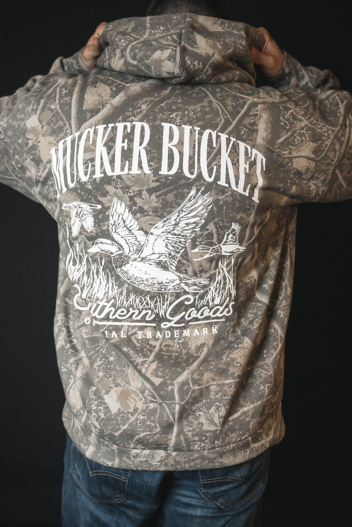 A person faces away in a Mallard Deadwood Camo Hoodie by Mucker Bucket, featuring stacking ducks, reeds, and a dog graphic on the back, plus Southern Goods and TRADEMARK.