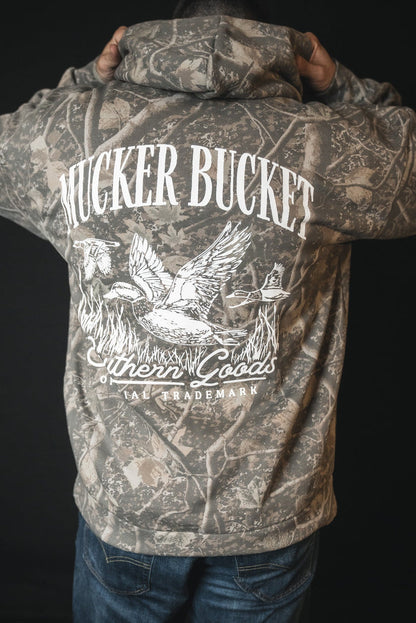 A person faces away in a Mallard Deadwood Camo Hoodie by Mucker Bucket, featuring stacking ducks, reeds, and a dog graphic on the back, plus Southern Goods and TRADEMARK.