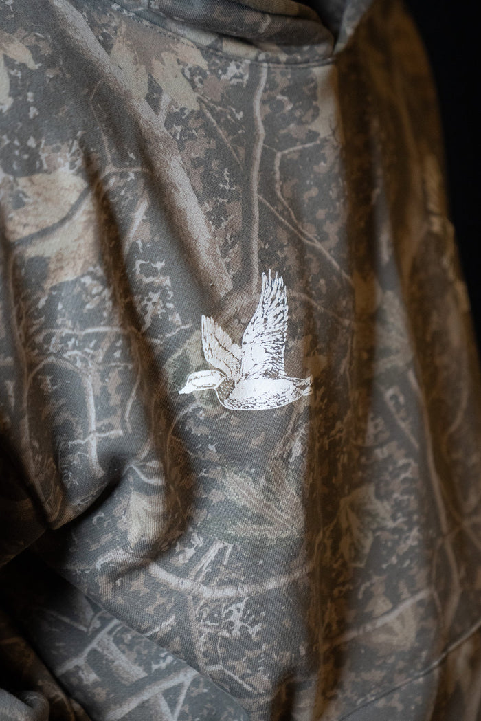 A close-up of the Mucker Bucket Mallard Deadwood Camo Hoodie, showcasing its trendy camouflage pattern and an embroidered white duck in flight on the back.
