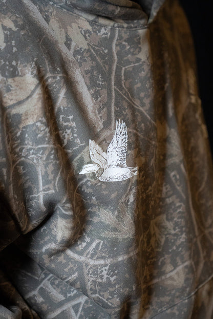 A close-up of the Mucker Bucket Mallard Deadwood Camo Hoodie, showcasing its trendy camouflage pattern and an embroidered white duck in flight on the back.
