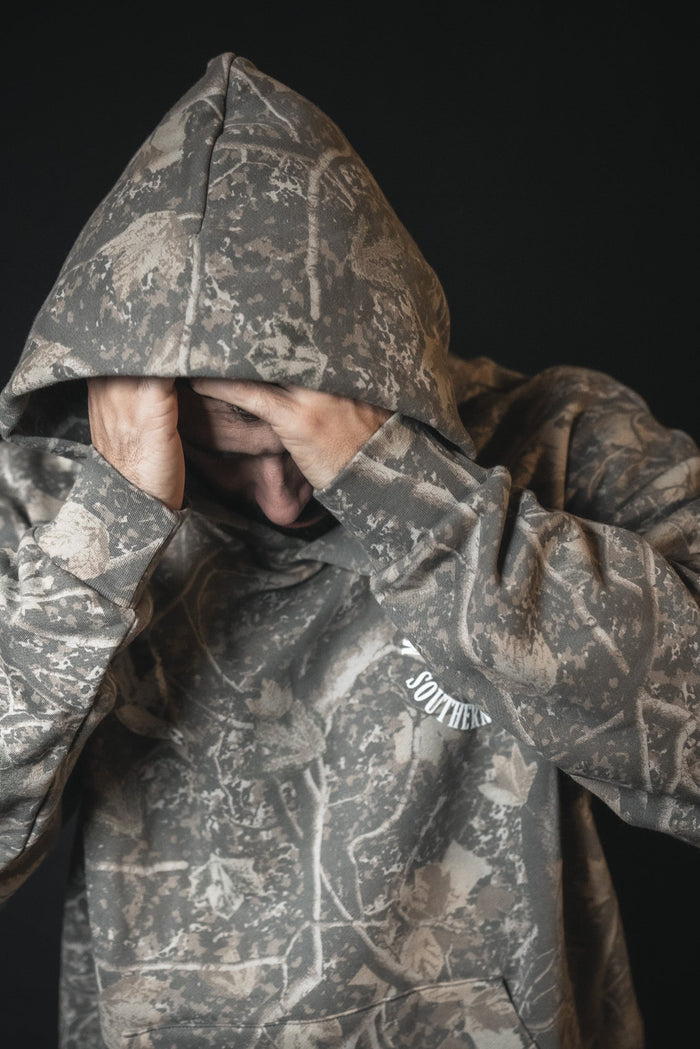 A person in a Mucker Bucket Mallard Deadwood Camo Hoodie with the hood up covers their face with both hands, standing against a dark background.