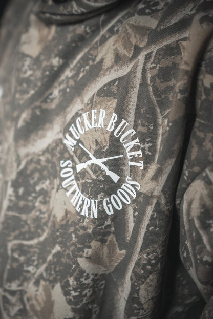 A close-up of the Mucker Bucket Mallard Deadwood Camo Hoodie featuring a white MUCKER BUCKET SOUTHERN GOODS logo and two crossed rifles—perfect to pair with your favorite premium relaxed fit styles.