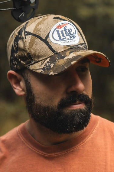 A bearded man in an orange shirt stands outdoors wearing a Mucker Bucket Mallard Life Deadwood Camo Bucket hat, looking slightly down and to the side. The green and brown-toned background is blurred.