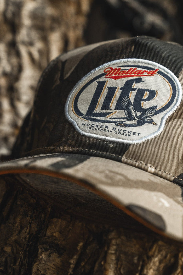The Mucker Bucket Mallard Life Deadwood Camo Bucket, featuring a flying duck patch, rests on a tree trunk outdoors.