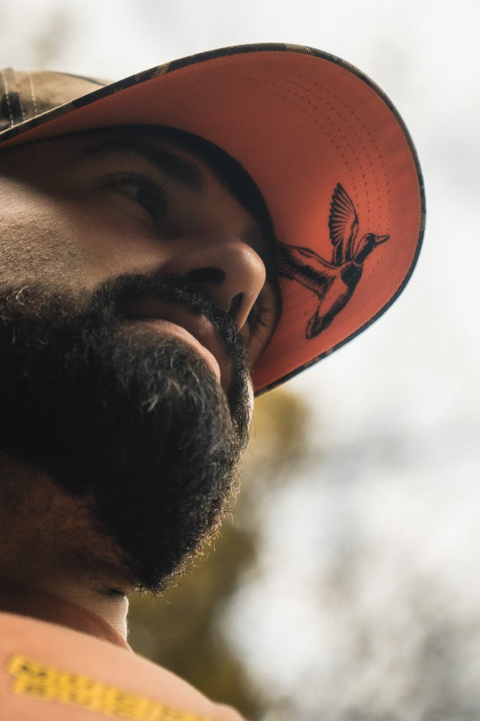A bearded person wearing the Mucker Bucket Mallard Life Deadwood Camo Bucket looks up toward the sky, with a blurred tree background enhancing the serene mallard life vibe.