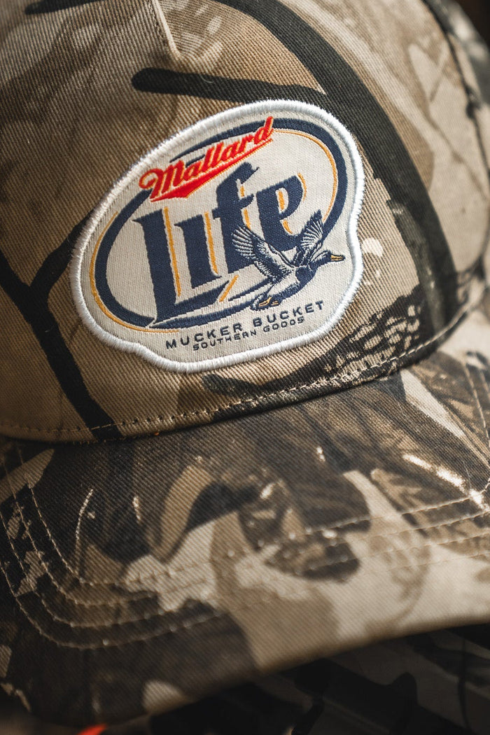 A close-up of the Mallard Life Deadwood Camo Bucket by Mucker Bucket, featuring a patch with a duck graphic and text, resting on a dark surface.