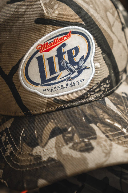 A close-up of the Mallard Life Deadwood Camo Bucket by Mucker Bucket, featuring a patch with a duck graphic and text, resting on a dark surface.