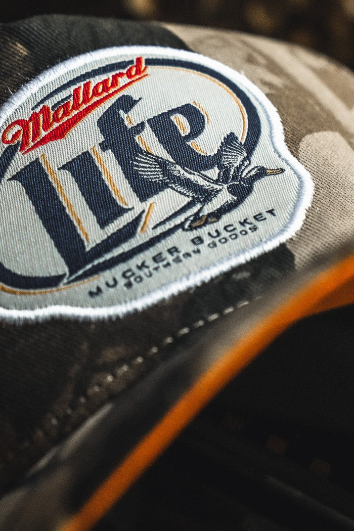 Close-up of the Mucker Bucket Mallard Life Deadwood Camo Bucket hat with a patch displaying "Mallard Life," a flying duck, and small text: Muck, Bucket, Guns, Mud, Dogs.