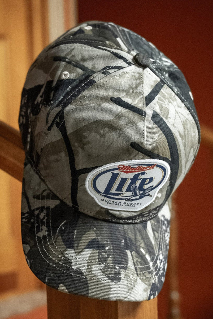 A Mucker Bucket Mallard Life Deadwood Camo Bucket hat hangs on a wooden banister indoors, with a wooden door and red wall in the background.