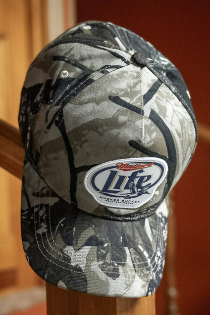 A Mucker Bucket Mallard Life Deadwood Camo Bucket hat hangs on a wooden banister indoors, with a wooden door and red wall in the background.
