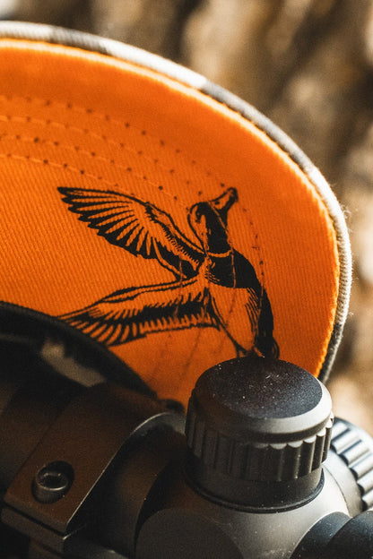 A close-up of the Mallard Life Deadwood Camo Bucket by Mucker Bucket, featuring a black flying duck design—perfect for mallard enthusiasts—with a blurred scope or lens in the foreground.
