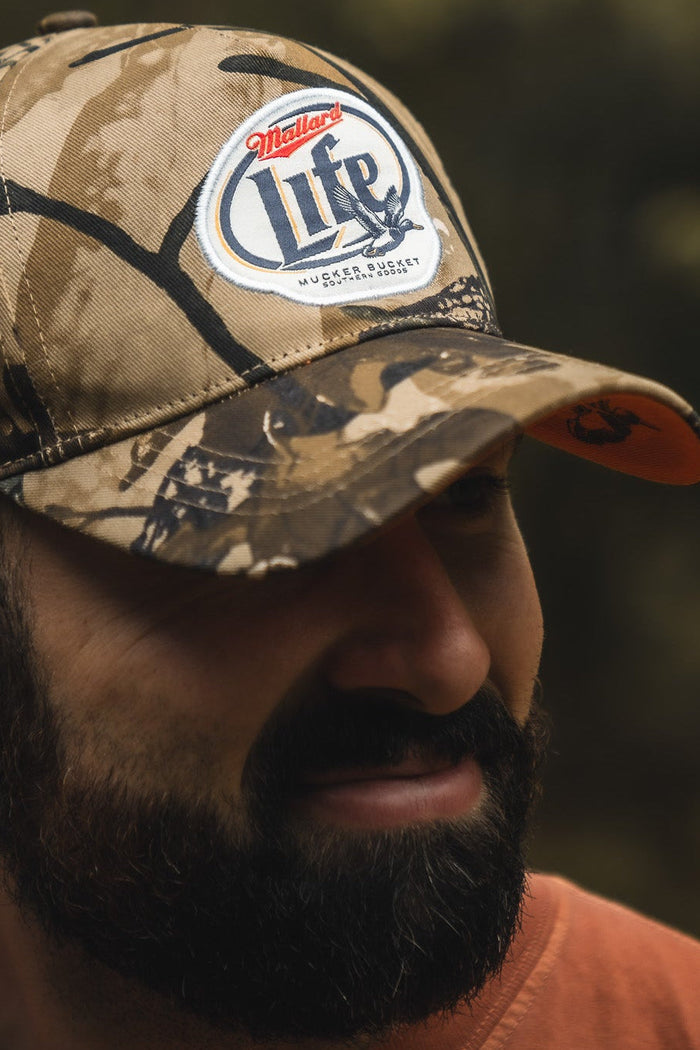 A man with a dark beard wears a Mucker Bucket Mallard Life Deadwood Camo Bucket hat. The blurred background in earthy tones hints at the true mallard life.