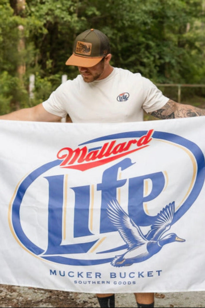 A man wearing a white t-shirt and trucker hat holds a Mucker Bucket Mallard Life Flag, featuring a flying duck logo and metal grommets, outdoors with trees in the background.