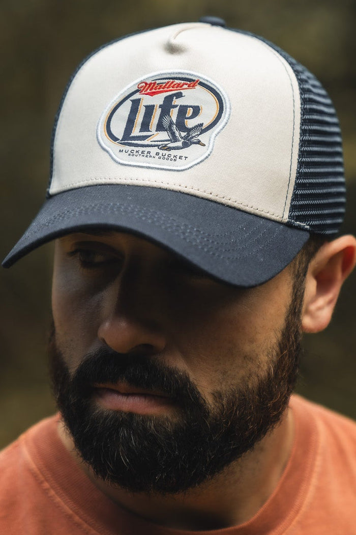 A bearded man in an orange shirt wears a Mucker Bucket Mallard Life Trucker Bucket hat with a mesh back, looking down with a neutral expression. The background is outdoors and blurred.