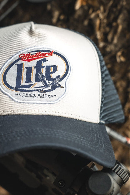 A close-up of the Mucker Bucket Mallard Life Trucker Bucket hat featuring a white front, navy bill, mesh back, and a patch with two ducks in flight and the text "Mallard Life" by Mucker Bucket.