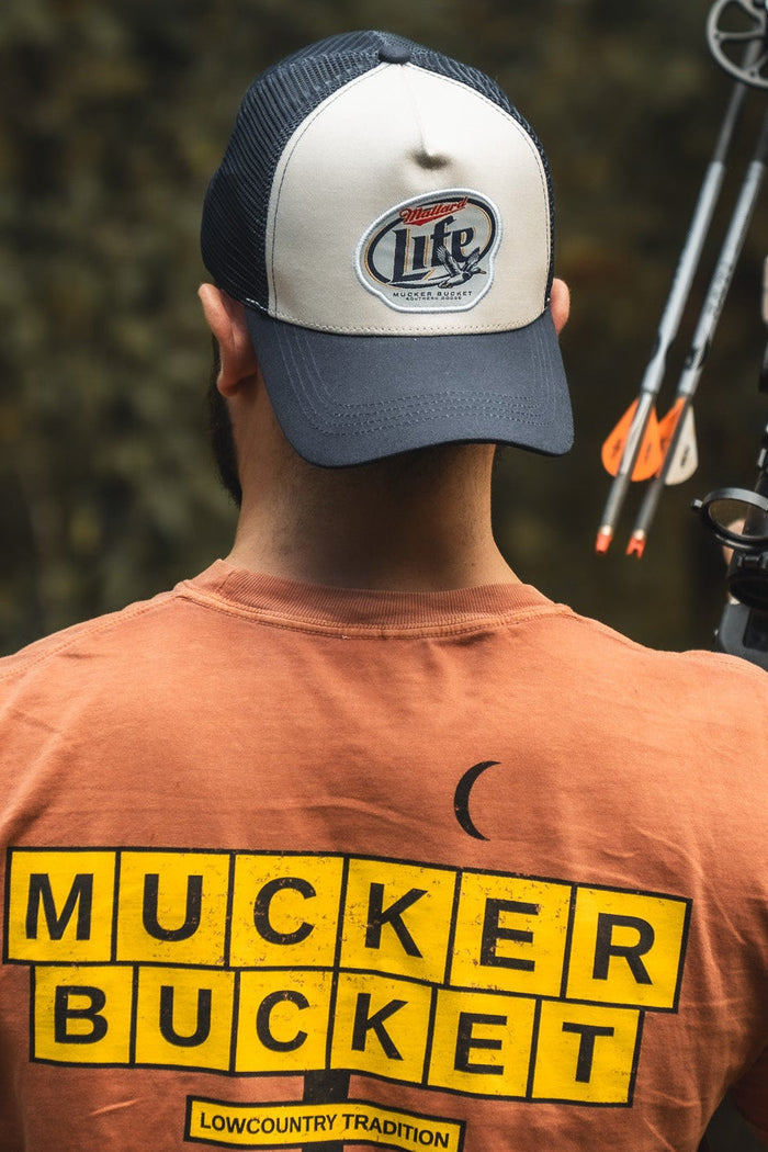 A person seen from behind wears an orange Mallard Life Trucker Bucket cap by Mucker Bucket and carries a compound bow with arrows over one shoulder.