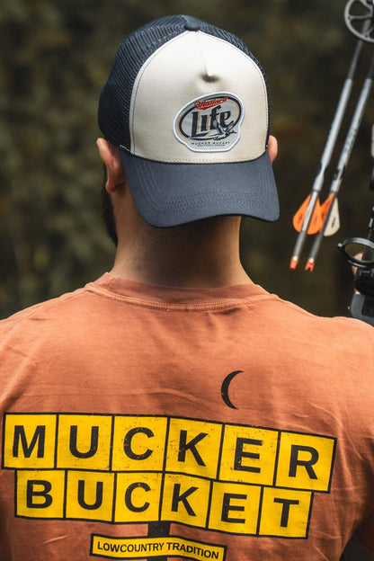A person seen from behind wears an orange Mallard Life Trucker Bucket cap by Mucker Bucket and carries a compound bow with arrows over one shoulder.