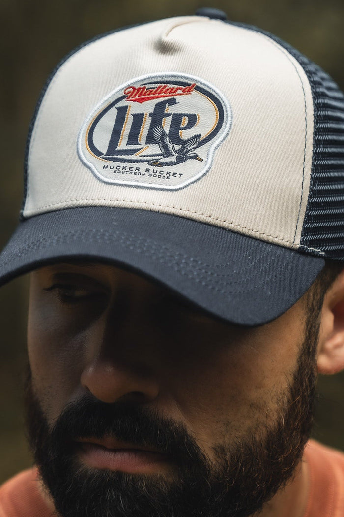 A bearded man wears a white and navy Mucker Bucket Mallard Life Trucker Bucket hat featuring a Mallard Life patch, mesh back, and trucker snapback. The blurred background highlights both the hat and his face.