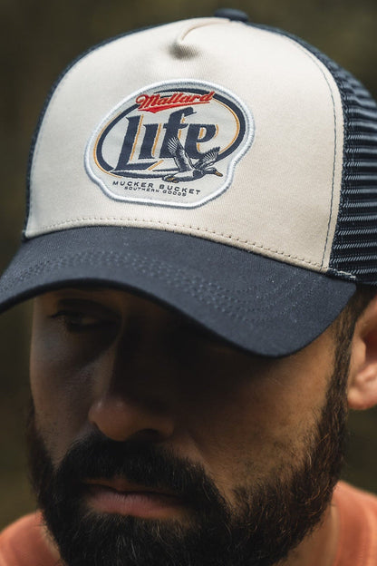 A bearded man wears a white and navy Mucker Bucket Mallard Life Trucker Bucket hat featuring a Mallard Life patch, mesh back, and trucker snapback. The blurred background highlights both the hat and his face.