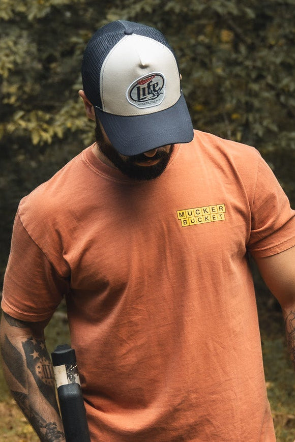 A tattooed man in an orange Mucker Bucket shirt and a Mallard Life Trucker Bucket snapback with mesh back stands outdoors holding a black object, surrounded by trees and greenery.