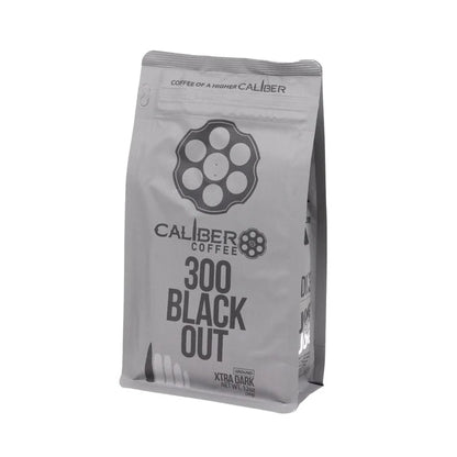 A gray coffee bag with Caliber Dark Roast 300 Blackout - Ground text and a revolver chamber design features Coffee of a higher caliber and Xtra Dark. Made from Specialty Grade, Single Origin South American beans, it stands upright. Brand: Caliber Coffee Company.