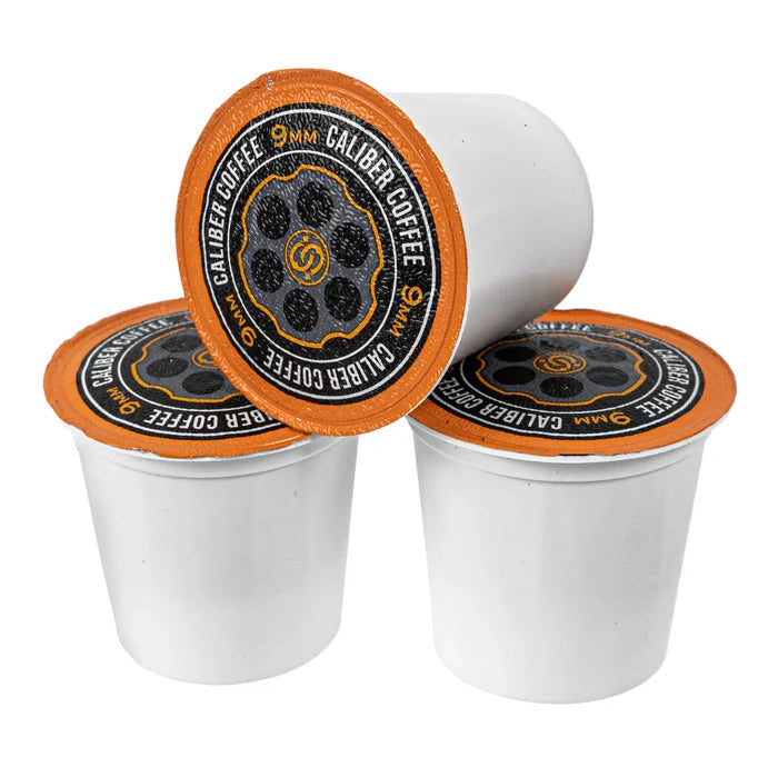 Three white Medium Roast 9MM Single-Serve Shells from Caliber Coffee Company, featuring orange lids with circular designs and text Caliber Coffee and 9MM, are stacked in a pyramid against a white background, perfect for a Colombian coffee experience.
