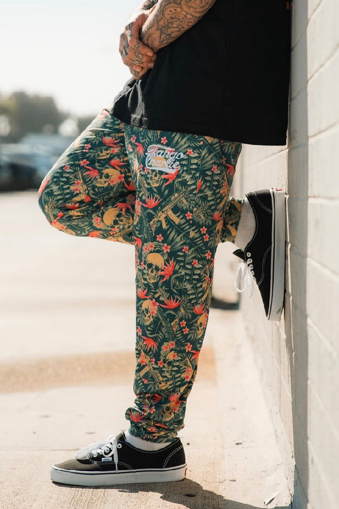 A tattooed person stands outdoors against a light brick wall, wearing Tango Charlie Apparel’s Men's Classic Premium Joggers | Aloha AR with a tropical pattern—perfect for running—paired with a black shirt and black sneakers. Cropped from the shoulders up.